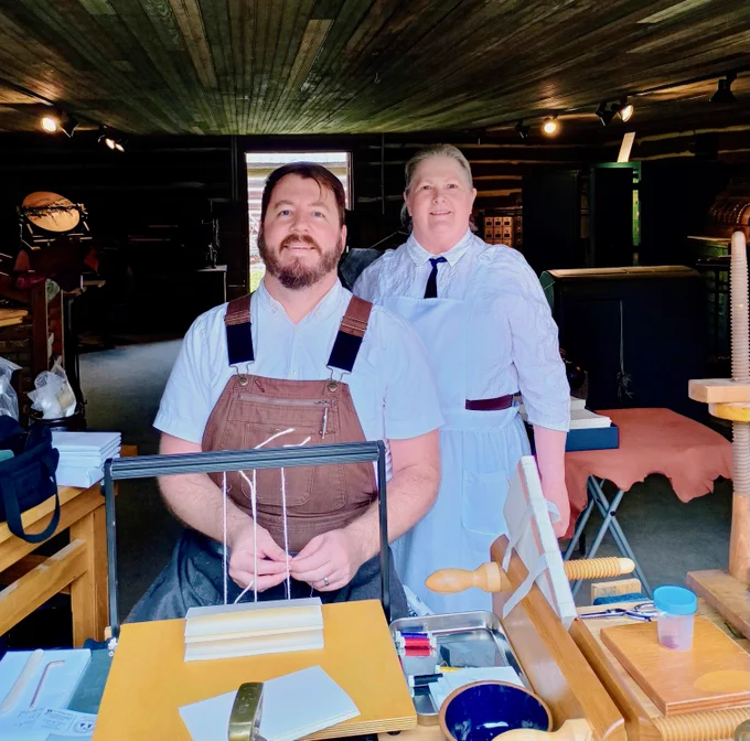 Two individuals are in a rustic, wood-paneled workshop. One is seated at a wooden table with bookbinding tools and materials, including paper and thread, while the other stands behind. They are both wearing period-style clothing. The setting suggests a historical or craft demonstration environment.