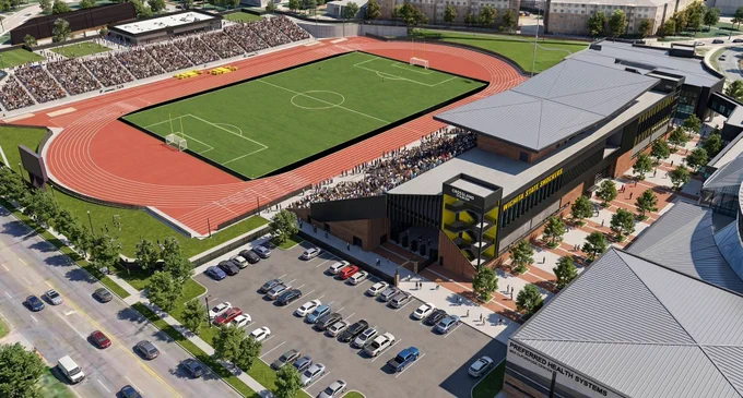 Aerial view of a sport complex. The facility includes a green field surrounded by a red running track. Stands full of spectators are present on one side. Adjacent to the field are parking lots and modern buildings. Streets and greenery surround the facility.