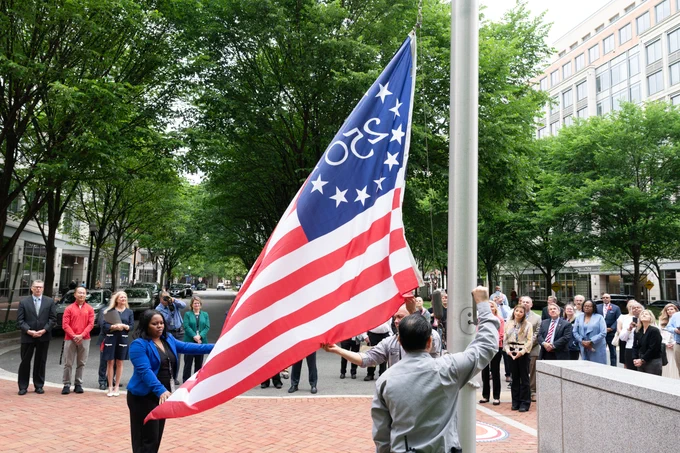 Group stands behind the America 250 flag as it is being raised by three people.