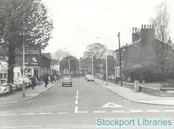 A street scene with parked cars, pedestrians, and terraced houses.