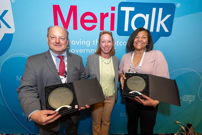 Two professionals holding MeriTalk awards while standing beside a woman in front of a MeriTalk branded backdrop.