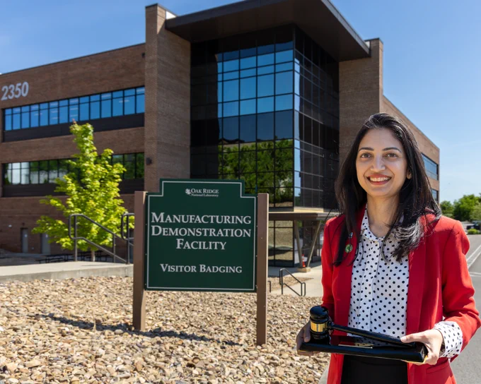 A woman stands outside the Oak Ridge National Laboratory's Manufacturing Demonstration Facility, holding a gavel. The building has large windows, and there's a sign for visitor badging.