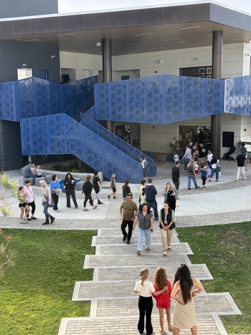 Families walk through a modern school courtyard with blue staircases during an evening campus event.