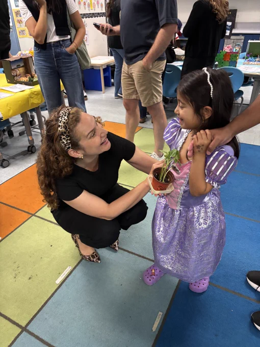 Teacher smiles while presenting a potted plant to a young student in a purple princess dress during classroom night.