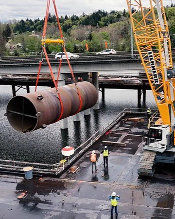 A crane is lifting a large cylindrical metal pipe over a body of water on a construction site. Workers in safety gear are guiding the operation. A bridge and greenery are visible in the background.