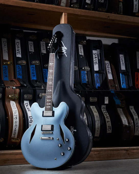 A light blue Epiphone DG-335 guitar stands leaning against its black guitar case. Behind it, there are several shelves filled with guitar cases labeled with different names and tags, some mentioning "Dave" and "Tour" referring to Dave Grohl of the Foo Fighters.