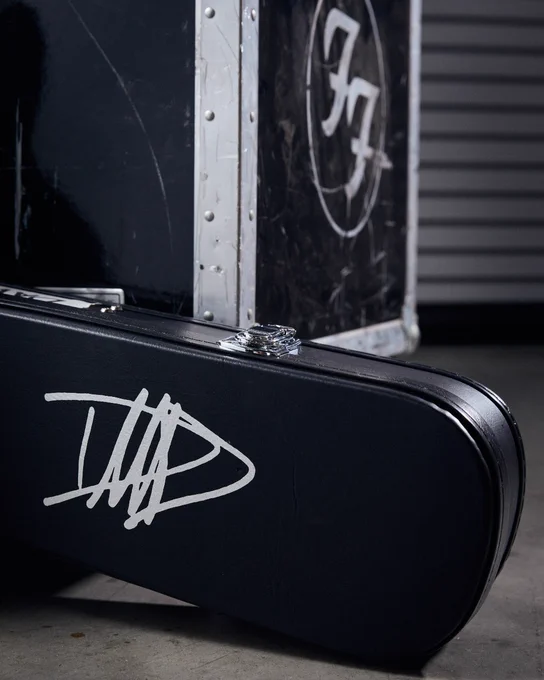A black guitar case with a white Dave Grohl signature rests against a black road case featuring the Foo Fighters logo. The background includes a metal shutter.