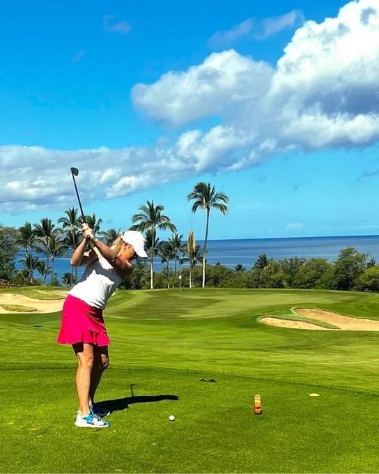 A person is playing golf on a lush green course with palm trees in the background. The ocean and a partly cloudy sky are visible beyond the course.