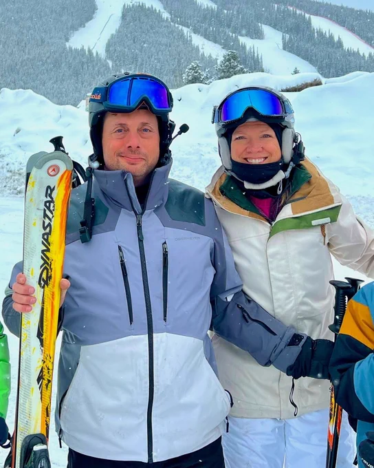 Two people dressed in ski gear, including helmets and goggles, stand side by side on a snowy slope. One is holding Dynastar skis. A snow-covered mountain is visible in the background.