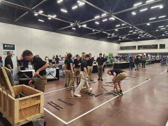 Members of the Michigan Tech Steel Bridge Team assemble sections of their steel bridge during the 2026 Student Steel Bridge Competition National Finals in a large indoor competition hall in El Paso, Texas. Team members in black Michigan Tech polos and khaki pants carry and position steel bridge components inside the marked construction area while other competitors and spectators are visible in the background.