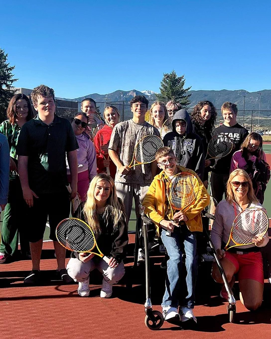 A group of people on a tennis court, smiling and holding rackets. One person is using a walker. They are outdoors with a view of mountains in the background and a clear blue sky. Some individuals are wearing sportswear, and a variety of racket brands are visible.