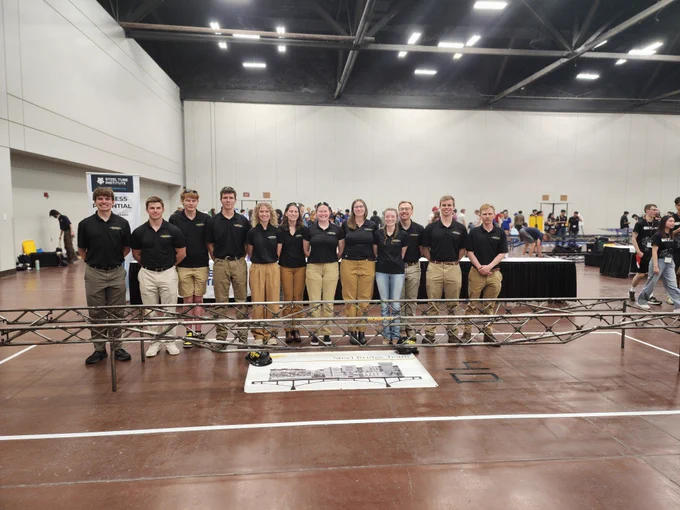 The Michigan Tech Steel Bridge Team poses behind their completed steel bridge at the 2026 Student Steel Bridge Competition National Finals. Team members stand shoulder to shoulder in matching black polos and khaki pants inside the competition venue with the bridge displayed prominently in the foreground.