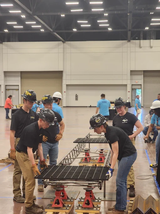Members of the Michigan Tech Steel Bridge Team prepare their bridge for structural loading during the national competition. Students wearing safety helmets and work gloves position deck panels onto the steel bridge while judges and volunteers observe nearby in the testing area.
