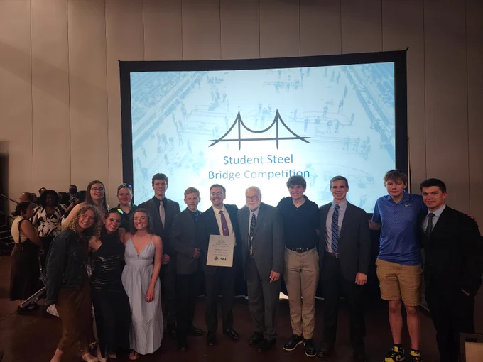 Members of the Michigan Tech Steel Bridge Team pose with competition officials during the awards banquet at the 2026 Student Steel Bridge Competition National Finals. The group stands in front of a large presentation screen displaying the Student Steel Bridge Competition logo while holding a competition award plaque.