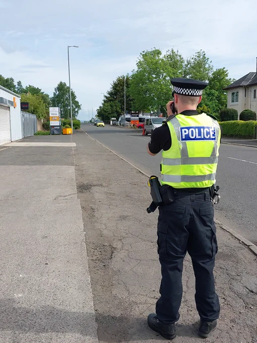 A police officer in uniform, wearing a high-visibility vest, is doing speed checks on a pavement.