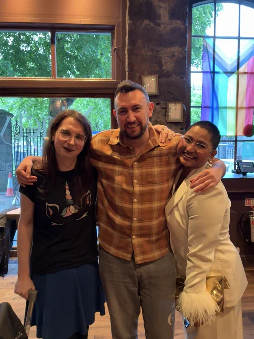 Left to right: Esme Houston (dressed in a black t-shirt with a trans flag coloured unicorn print and a dark blue skirt), Guy Ingerson (dressed in an orange checked shirt and grey jeans), and Sai Shraddha Suresh Viswanathan (dressed in a white pant suit with feathered sleeves).

A progress pride flag hangs from a window behind them.