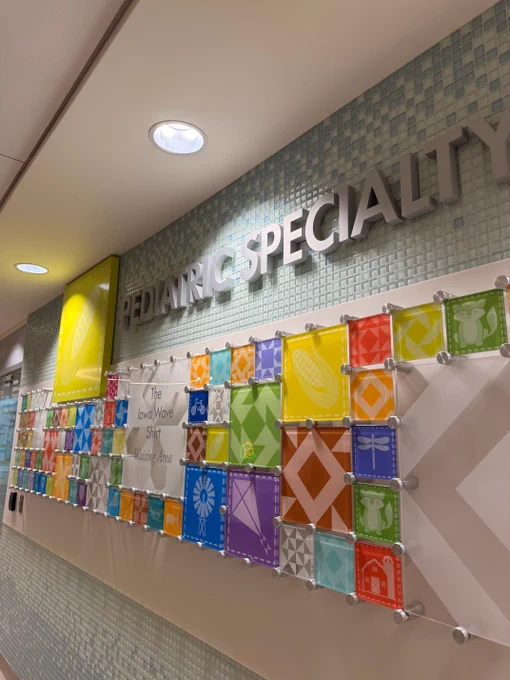 Wall in a pediatric specialty area featuring colorful square panels with various icons like a butterfly, corn, and windmill. The display includes a sign reading "The Iowa Wave Shirt Waiting Area" on a light blue tiled background.