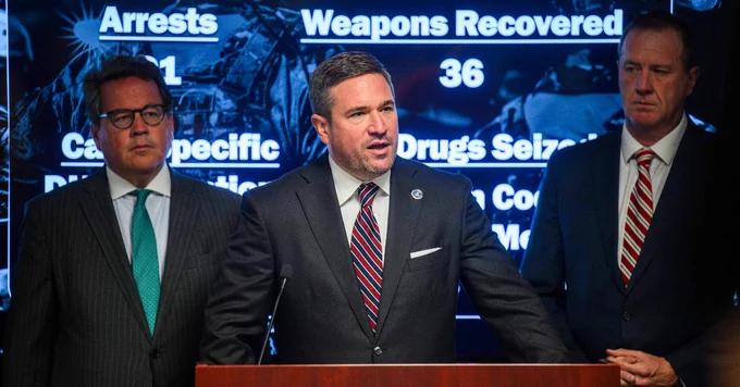 Three men in suits stand at a podium during a press briefing with crime statistics displayed on a screen behind them.