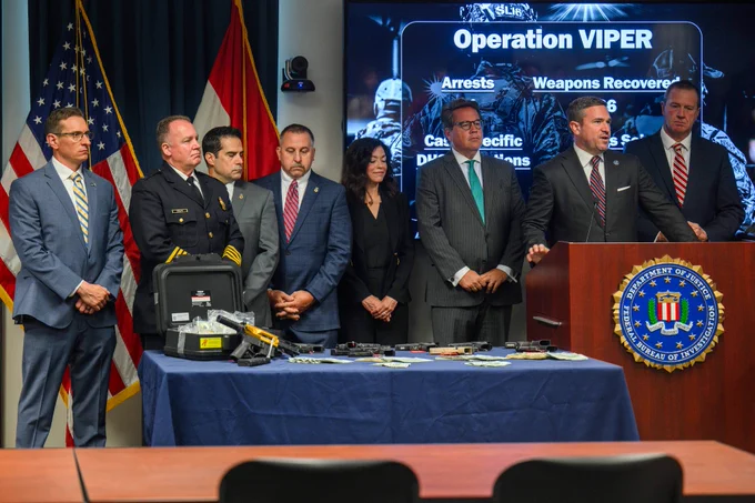 Group of law enforcement officials standing behind a table displaying seized weapons and evidence during a press briefing on Operation VIPER.