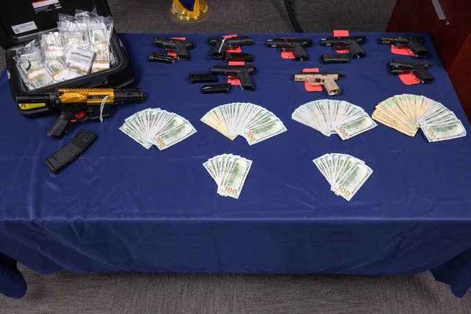 Table displaying multiple firearms, stacks of cash in various denominations, and a suitcase filled with money bundles on a blue cloth.