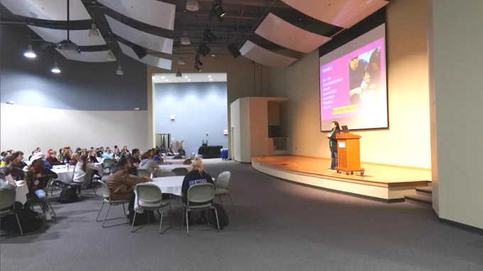 A large conference room with an audience seated at round tables, listening to a speaker on stage. The speaker is presenting a slide with text and images. The room is well-lit with high ceilings and sound panels.