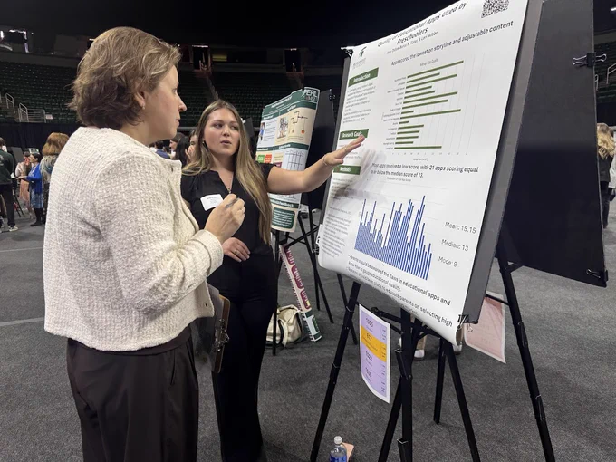 A student presents research titled \"Quality of Educational Apps Used by Preschoolers\" to a woman. The poster includes bar graphs showing app quality ratings and notes that \"Apps scored the lowest on storyline and adjustable content.\"