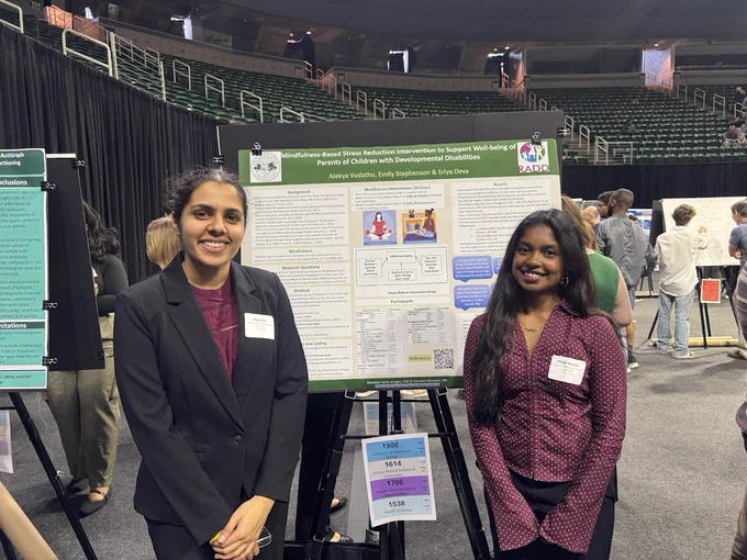 Two South Asian women stand next to a poster titled \"Mindfulness-Based Stress Reduction Intervention to Support Well-being of Parents of Children with Developmental Disabilities\" by Alekya Vudathu, Emily Stephenson & Sriya Deva. In the foreground, other posters and people are visible.