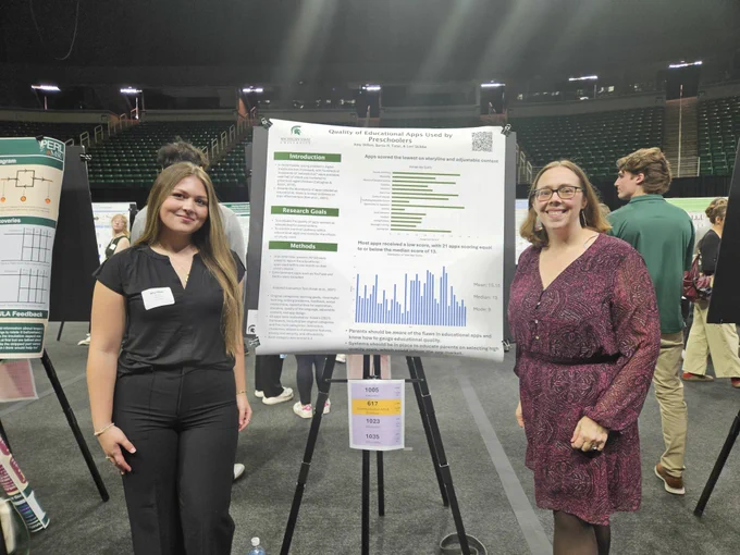 Two women stand beside a poster titled \"Quality of Educational Apps Used by Preschoolers\" by Amy Dillon, Burcu H. Tatar, & Lori Skibbe. The poster includes graphs and text about app quality. On the poster are texts 1005@number, 617@number, 1023@number and 1035@number.