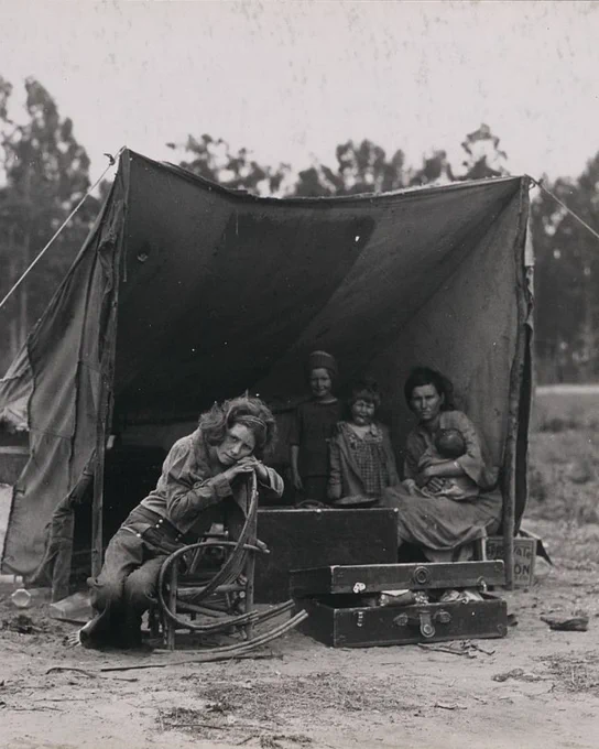 An image in the same series as Dorothea Lange's famous "Migrant Mother" photograph features the same mother holding a baby, but from a wider angle that shows the entire lean-to tent structure she sits under, along with three other children standing or sitting near her. In this image, all of the subjects appear to be looking right at the camera.