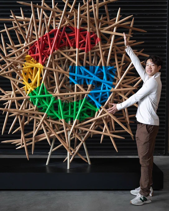 Daniel poses next to an abstract wooden stick sculpture on a wall containing a colorful Google "G" logo.
