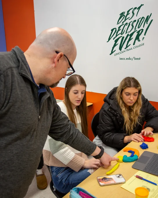 Teacher pointing at a yellow clay shape that is part of a model of the brain while two students watch attentively in a bright, modern classroom.