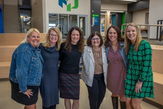 Group of six women standing closely together inside a modern office space with wooden steps and glass walls.