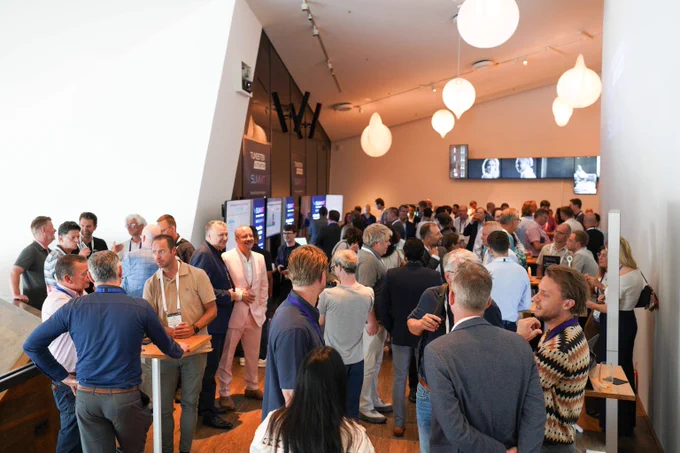 A busy networking event with professionals engaged in conversations under modern pendant lights in a spacious, well-lit room.