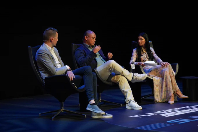 Three people seated on stage chairs engaged in a panel discussion under focused lighting against a dark background.