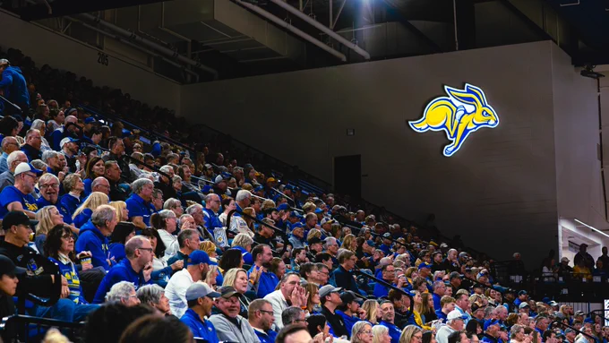 A wide view of the arena seating shows SDSU fans wearing blue and yellow apparel filling the stands, with many clapping and watching the game, while a large illuminated Jackrabbit logo is displayed on the wall above the seating area.