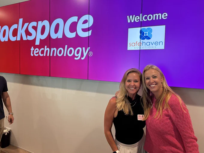 Two individuals smile in front of a large display screen showing the Rackspace Technology logo on a purple background. The screen also features the Safe Haven Solutions logo with a "Welcome" message.