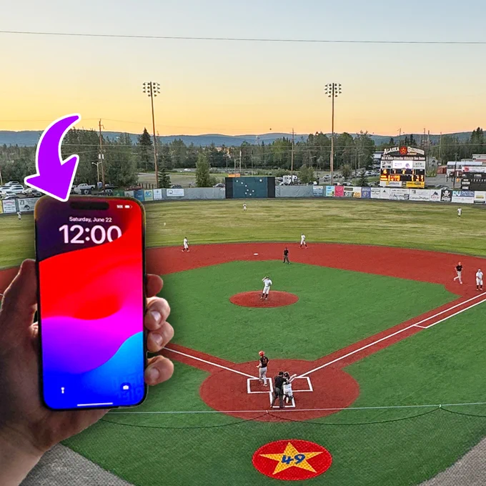 A baseball field looking outward to the surrounding skyline. A hand holding a cellphone is overlaid on top of the lower left portion. A purple arrow is pointing to the “12:00” on the phone screen.