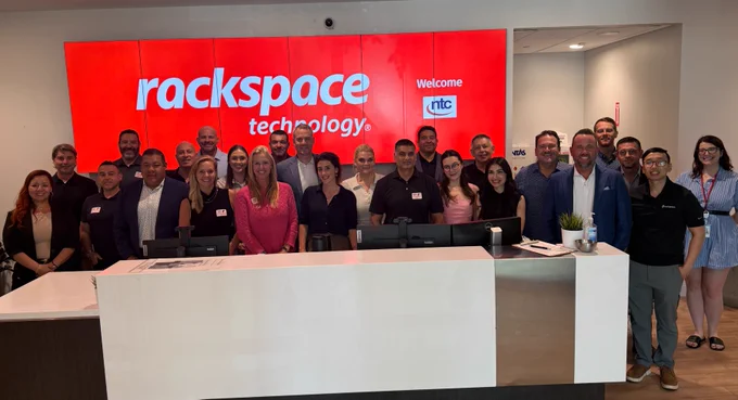 A group of people stand together in front of a reception area at Rackspace Technology, with a large red sign displaying the company name on the wall behind them. They are smiling and facing the camera.