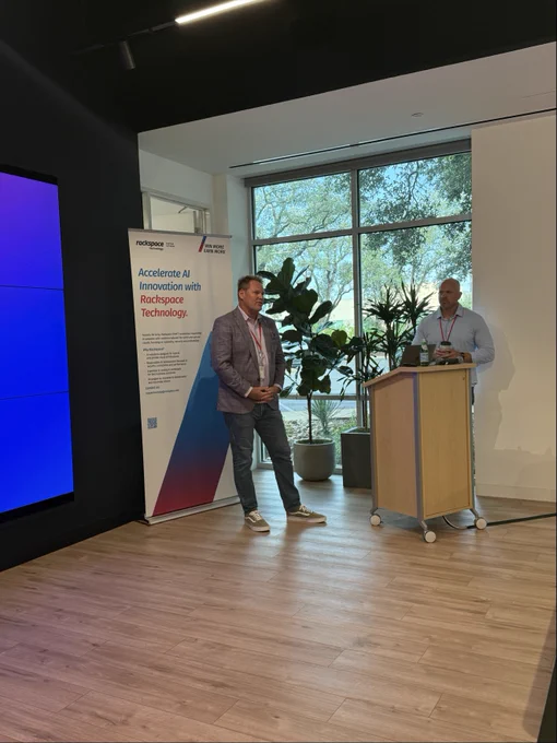 Two individuals are in a room with wooden flooring and large windows. One is standing beside a vertical banner displaying "Rackspace Technology." The other is at a podium with a laptop. Potted plants are in the background.