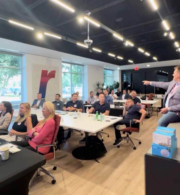 A group of people are seated in a modern, well-lit conference room with large windows. They are facing a presenter at the front who is gesturing towards a screen. There are several tables with drinks and notepads. A logo resembling the letter "R" is displayed on the wall.