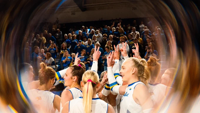 SDSU volleyball players wearing white uniforms gather in a tight huddle with hands raised together, viewed through a circular foreground effect, while a crowd of fans in blue and gold stands in the arena seating behind them.