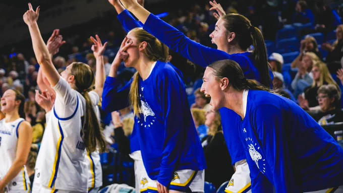 SDSU women's basketball players along the bench wear blue warm‑up shirts and white uniforms, leaning forward and raising their arms while pointing toward the court during a game, with spectators seated in the arena behind them.