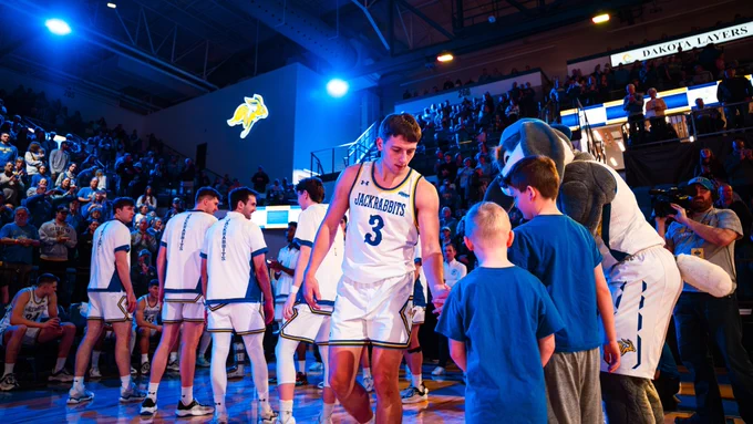An SDSU men's basketball player wearing a white “Jackrabbits” jersey with the number 3 walks along the court and interacts with fans at the sideline, while teammates in white uniforms stand nearby and the Jackrabbits mascot engages with children, with a full arena and a lit Jackrabbit logo visible in the background.