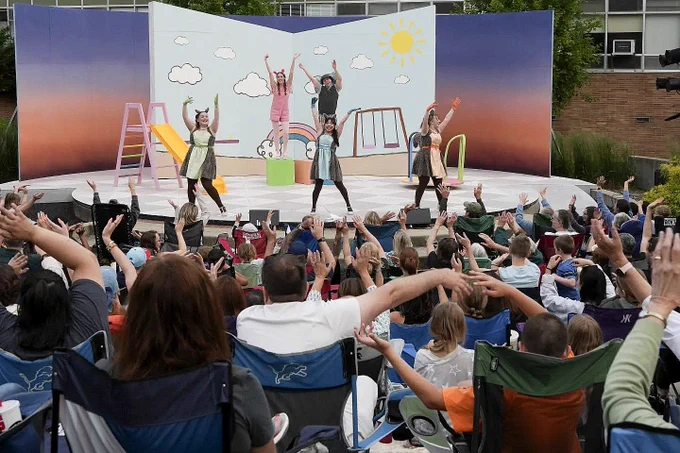 An energetic outdoor theatrical performance takes place on a stage featuring a backdrop shaped like an open, illustrated children's book. In the foreground, an audience seated in lawn chairs raises their arms to mirror the choreography of the actors on stage.