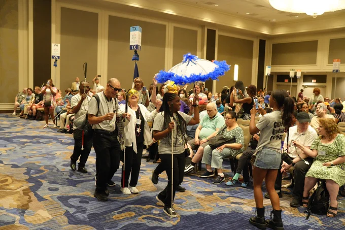 NFB of New Orleans Chapter members dance and celebrate as they follow the band through the convention hall. They wear traditional Mardi Gras items. 