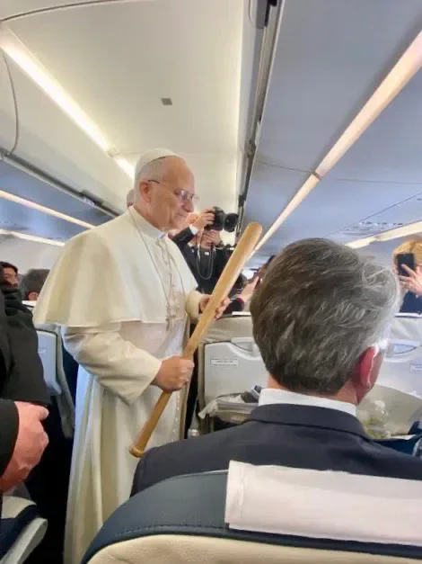 Pope Leo standing up in an airplane aisle holding a baseball bat 