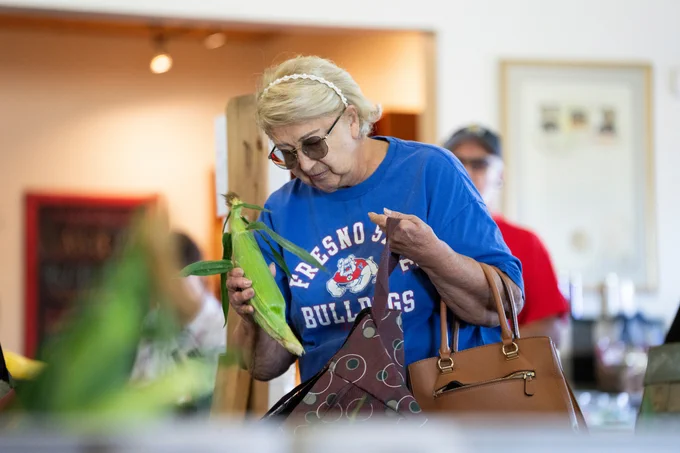A person in a blue Fresno State Bulldogs shirt examines a corn cob at a market. They have a handbag over their shoulder and wear sunglasses and a headband.
