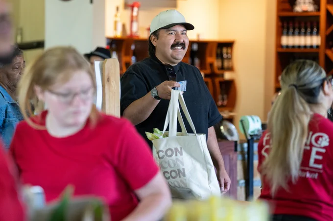 Person smiling while holding a shopping bag indoors. Individuals in red shirts are visible in the foreground, with "Fresno State" printed on one shirt.