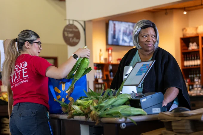 A person wearing a red Fresno State T-shirt is placing corn cobs into a blue Walmart shopping bag at a market checkout counter. Another person behind the counter is operating the register, smiling. In the background, shelves display various bottles and a wall-mounted TV is visible.
