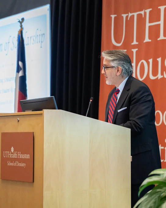 A person stands at a wooden podium giving a speech. The podium displays the UTHealth Houston School of Dentistry logo, and a large orange backdrop with the same branding fills the background. To the left, a projection screen shows the title “Celebration of Scholarship,” and the Texas flag stands nearby.